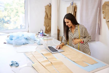 Foto de Mujer embarazada está en el trabajo en un estudio de costura, ella utiliza un ordenador portátil y patrones en su trabajo - Imagen libre de derechos