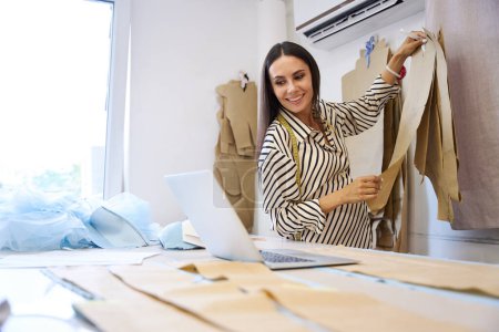 Téléchargez les photos : Femme enceinte sur le lieu de travail dans un atelier de couture, elle utilise un ordinateur portable et des motifs dans son travail - en image libre de droit