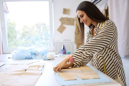 Photo for Pregnant craftswoman is at her workplace in a sewing studio, she lays out patterns on fabric - Royalty Free Image