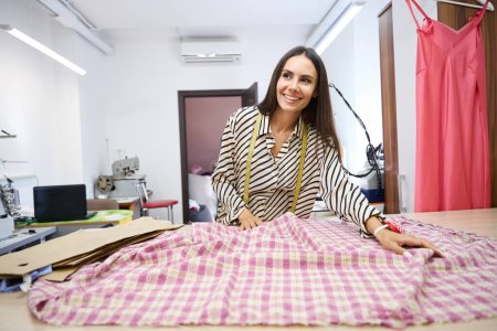 Téléchargez les photos : Femme souriante pose le tissu à carreaux sur la table de coupe, a un ruban à mesurer autour de son cou - en image libre de droit