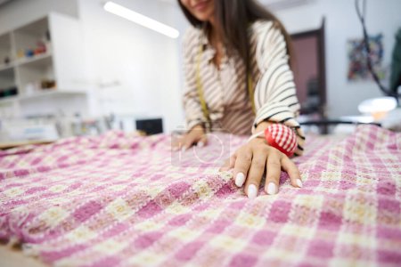 Photo for Designer lays out plaid fabric on the cutting table, she has a pincushion on her hand - Royalty Free Image