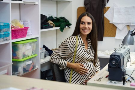Téléchargez les photos : Une femme enceinte souriante s'assoit sur son lieu de travail à une machine à coudre, dans un atelier d'équipement moderne - en image libre de droit