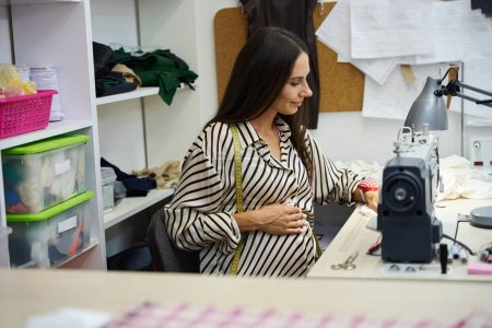 Foto de Smiling embarazada se sienta en su lugar de trabajo en una máquina de coser, en un taller con equipos modernos - Imagen libre de derechos