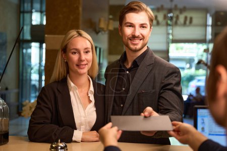 Photo for Amused businessman and businesswoman standing near reception desk and man giving black envelope to woman administrator - Royalty Free Image