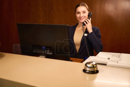 Téléchargez les photos : Accueil administrateur parler au téléphone tout en se tenant devant l'ordinateur à la réception de l'hôtel - en image libre de droit