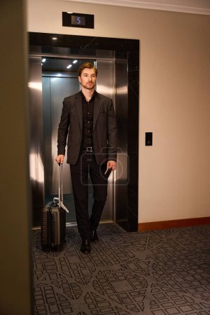 Foto de Tranquilo joven caminando por el ascensor en el hotel mientras sostiene la maleta en una mano y documentos en otra - Imagen libre de derechos