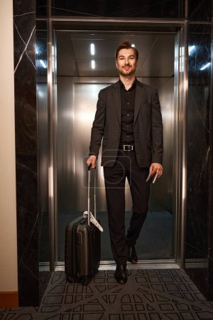 Téléchargez les photos : Photo pleine longueur de valise à roulettes homme d'affaires lors de la sortie de l'ascenseur à l'hôtel - en image libre de droit
