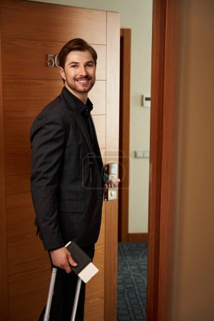 Foto de Joven hombre sonriente entrando en la habitación del hotel mientras sostiene documentos y equipaje en la mano - Imagen libre de derechos