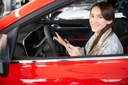 Foto de Feliz joven sonriente mujer sentada en un automóvil nuevo disfrutando de su compra en concesionario - Imagen libre de derechos
