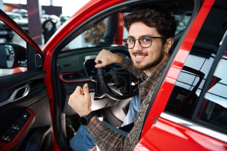 Foto de Atractivo hombre feliz comprar coche nuevo satisfecho de modelo que muestra como gesto sentado en auto en concesionario - Imagen libre de derechos