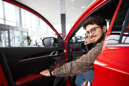 Téléchargez les photos : Beau homme heureux assis dans sa nouvelle cabine d'automobile tester la nouvelle voiture chez le concessionnaire - en image libre de droit