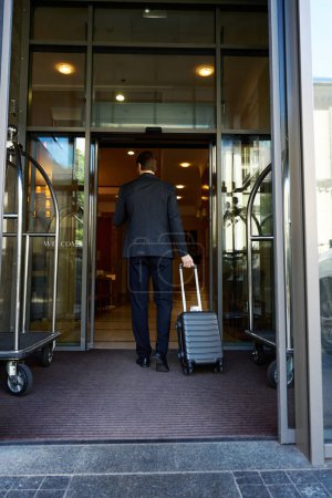 Téléchargez les photos : Vue de dos de l'homme d'affaires en costume tenant sac chariot par poignée à la main tout en entrant dans l'hôtel - en image libre de droit