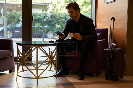Photo for Young man sitting on armchair at lobby bar while watching something on smartphone during coffeetime - Royalty Free Image