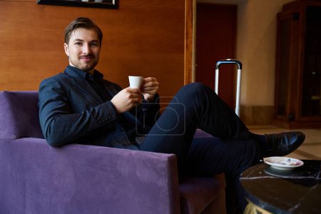 Photo for Close up photo of smiling guy sitting on armchair at hotel lobby bar and having coffee time - Royalty Free Image