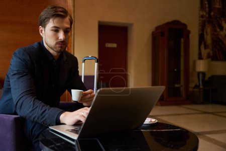 Foto de Hombre joven sentado en el sillón en la mesa en el bar del vestíbulo mientras trabaja en el ordenador portátil y beber café - Imagen libre de derechos
