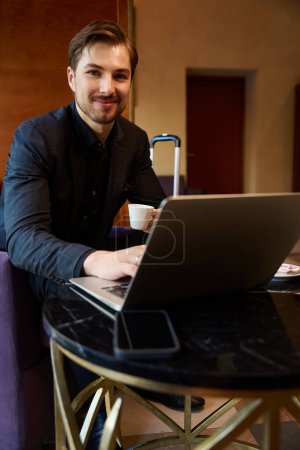 Foto de Foto de cerca del joven empresario alegre sentado en el vestíbulo del hotel mientras trabajaba en el ordenador portátil y bebía café - Imagen libre de derechos
