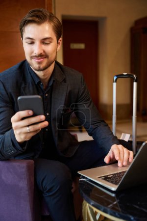 Foto de Imagen de cerca del hombre sonriente sentado en el vestíbulo del hotel mientras se comunica en línea por teléfono y trabaja en la computadora portátil - Imagen libre de derechos