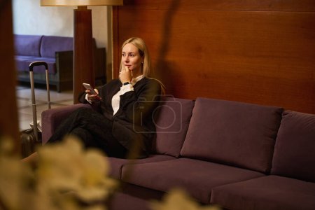 Téléchargez les photos : Femme satisfaite assise sur le canapé dans le hall tout en pensant à quoi regarder au téléphone - en image libre de droit