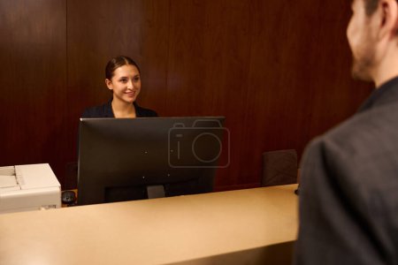 Téléchargez les photos : Administratrice sympathique debout à la réception devant moniteur et accueillant un visiteur à l'hôtel - en image libre de droit