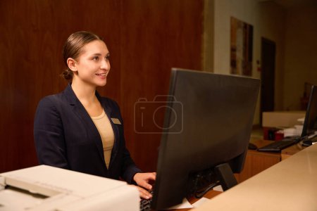 Foto de Recepcionista alegre saludando a alguien mientras está de pie frente a la computadora en el mostrador de recepción - Imagen libre de derechos