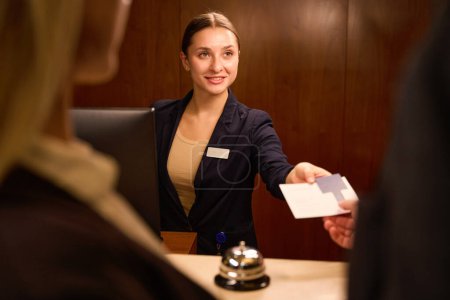 Téléchargez les photos : Photo recadrée d'une jeune réceptionniste joyeuse debout à la réception tout en donnant une carte-clé en plastique au visiteur de l'hôtel - en image libre de droit