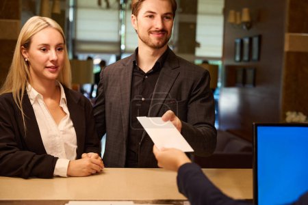 Téléchargez les photos : Jeune homme et jeune femme debout à la réception de l'hôtel tandis qu'un homme prend l'enveloppe en papier de la main de l'administrateur - en image libre de droit
