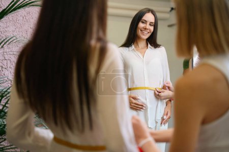 Foto de Embarazada cliente está teniendo un ajuste en un taller de costura, las mujeres se colocan delante de un gran espejo - Imagen libre de derechos