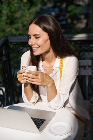 Téléchargez les photos : Jeune femme dégustant une tasse de café sur un charmant balcon avec un ruban à mesurer autour du cou - en image libre de droit