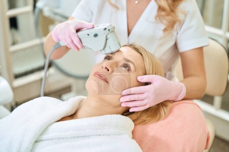 Photo for Cropped female cosmetologist doing laser depilation with laser hair removal machine of forehead of adult blonde caucasian woman in blurred beauty salon. Concept of face skin care - Royalty Free Image