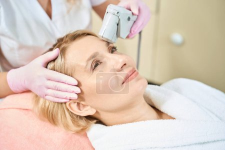 Photo for Partial female beautician doing laser depilation with laser hair removal machine of forehead of smiling adult caucasian woman in blurred beauty salon. Concept of face skin care - Royalty Free Image
