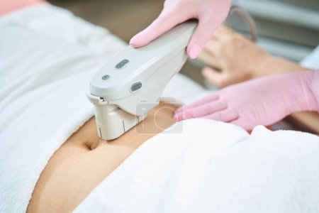 Photo for Cropped image of female beautician doing laser depilation with laser hair removal machine of woman belly in blurred beauty salon. Concept of body skin care - Royalty Free Image