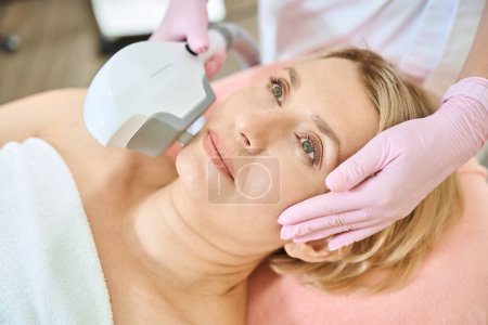 Photo for Partial female beautician wearing latex gloves doing smas-lifting with IPL photorejuvenation device of face of adult caucasian woman in blurred beauty salon. Concept of face skin care and rejuvenation - Royalty Free Image