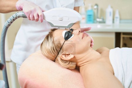 Photo for Side view of partial female cosmetologist doing smas-lifting with IPL photorejuvenation device of forehead of adult woman wearing glasses in blurred beauty salon. Face skin care and rejuvenation - Royalty Free Image