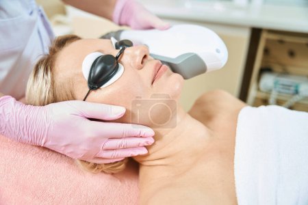 Photo for Cropped female cosmetologist doing smas-lifting with IPL photorejuvenation device of face of adult woman wearing glasses in blurred beauty salon. Concept of face skin care and rejuvenation - Royalty Free Image