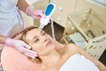Photo for Cropped female cosmetologist doing ultrasonic cleaning of face skin with IPL photorejuvenation device of adult caucasian woman on couch in blurred beauty salon. Face skin care and rejuvenation - Royalty Free Image