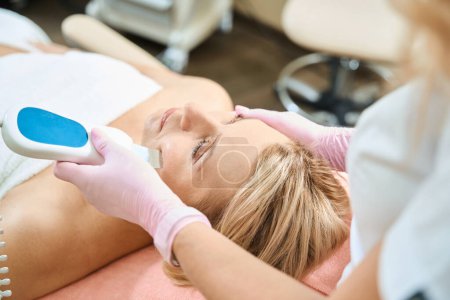 Photo for Partial female beautician doing ultrasonic cleaning of face skin with IPL photorejuvenation device of adult caucasian woman in blurred beauty salon. Concept of face skin care and rejuvenation - Royalty Free Image