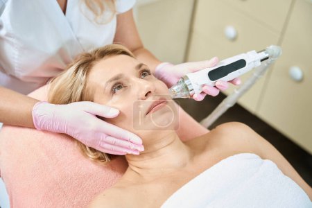 Photo for Cropped female cosmetologist doing microneedle rf lifting of face skin with IPL photorejuvenation device of adult caucasian woman in blurred beauty salon. Concept of face skin care and rejuvenation - Royalty Free Image