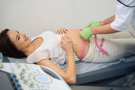 Téléchargez les photos : Gynécologue femelle cultivée mesurant le ventre avec du ruban adhésif de jeune femme enceinte caucasienne à la clinique. Concept d'examen de grossesse - en image libre de droit