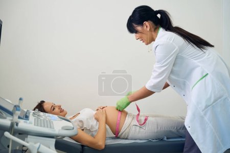 Téléchargez les photos : Vue latérale du gynécologue femelle mesurant le ventre avec du ruban adhésif de jeune femme enceinte caucasienne souriante à la clinique. Concept d'examen de grossesse - en image libre de droit
