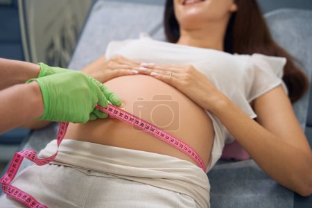 Téléchargez les photos : Image recadrée de gynécologue femelle mesurant le ventre avec du ruban adhésif de la femme enceinte à la clinique. Concept d'examen de grossesse - en image libre de droit