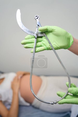 Téléchargez les photos : Concentration sélective de la jauge du bassin obstétricale dans les mains de gynécologue féminine avec fond de femme enceinte floue sur le canapé médical à la clinique. Concept d'examen de grossesse - en image libre de droit