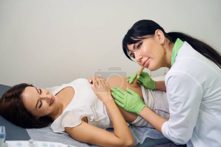 Téléchargez les photos : Femme gynécologue européenne concentrée adulte écoutant le ventre de la jeune femme enceinte caucasienne souriante en clinique. Concept d'examen de grossesse - en image libre de droit