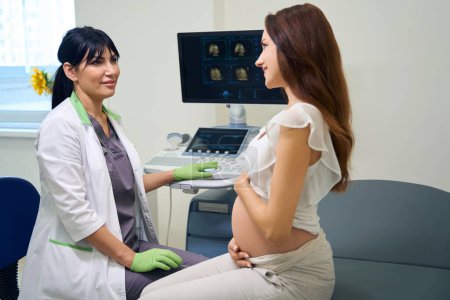 Téléchargez les photos : Gynécologue européenne adulte et jeune femme enceinte caucasienne se regardant lors d'un examen échographique en clinique. Concept d'examen de grossesse - en image libre de droit