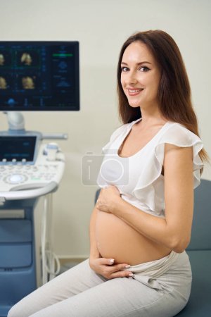 Photo for Young smiling caucasian pregnant woman looking at camera sitting on medical couch in ultrasound room in clinic. Concept of pregnancy examination - Royalty Free Image