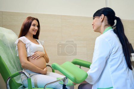 Téléchargez les photos : Gynécologue européenne adulte examinant une jeune femme enceinte caucasienne souriante sur un canapé médical à la clinique. Concept d'examen de grossesse - en image libre de droit
