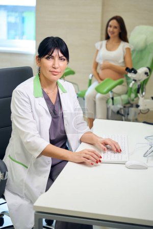 Téléchargez les photos : Gynécologue européenne adulte regardant la caméra en tapant sur l'ordinateur et en examinant femme enceinte souriante floue sur le canapé médical à la clinique. Concept d'examen de grossesse - en image libre de droit