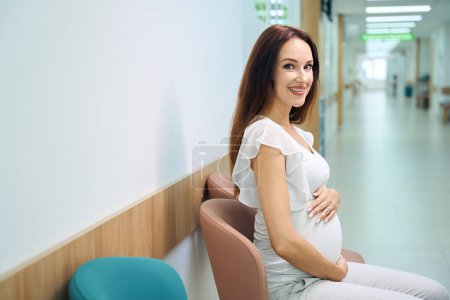 Téléchargez les photos : Jeune femme enceinte européenne souriante assise sur une chaise et regardant la caméra en attendant la consultation dans le hall de la clinique. Concept d'examen de grossesse - en image libre de droit