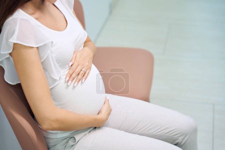Téléchargez les photos : Image partielle d'une femme enceinte assise sur une chaise et se touchant le ventre en attendant la consultation dans le hall de la clinique. Concept d'examen de grossesse - en image libre de droit