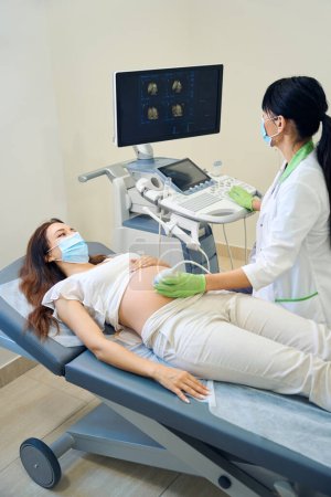 Photo for Adult european female gynecologist watching on monitor during doing ultrasound examination of young caucasian pregnant woman in clinic. Females wearing medical masks. Concept of pregnancy examination - Royalty Free Image