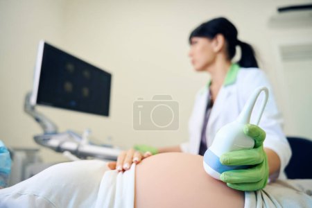 Foto de Enfoque selectivo del vientre de la mujer embarazada parcial durante ginecólogo femenino borroso haciendo un examen de ultrasonido en la clínica. Concepto de examen de embarazo - Imagen libre de derechos